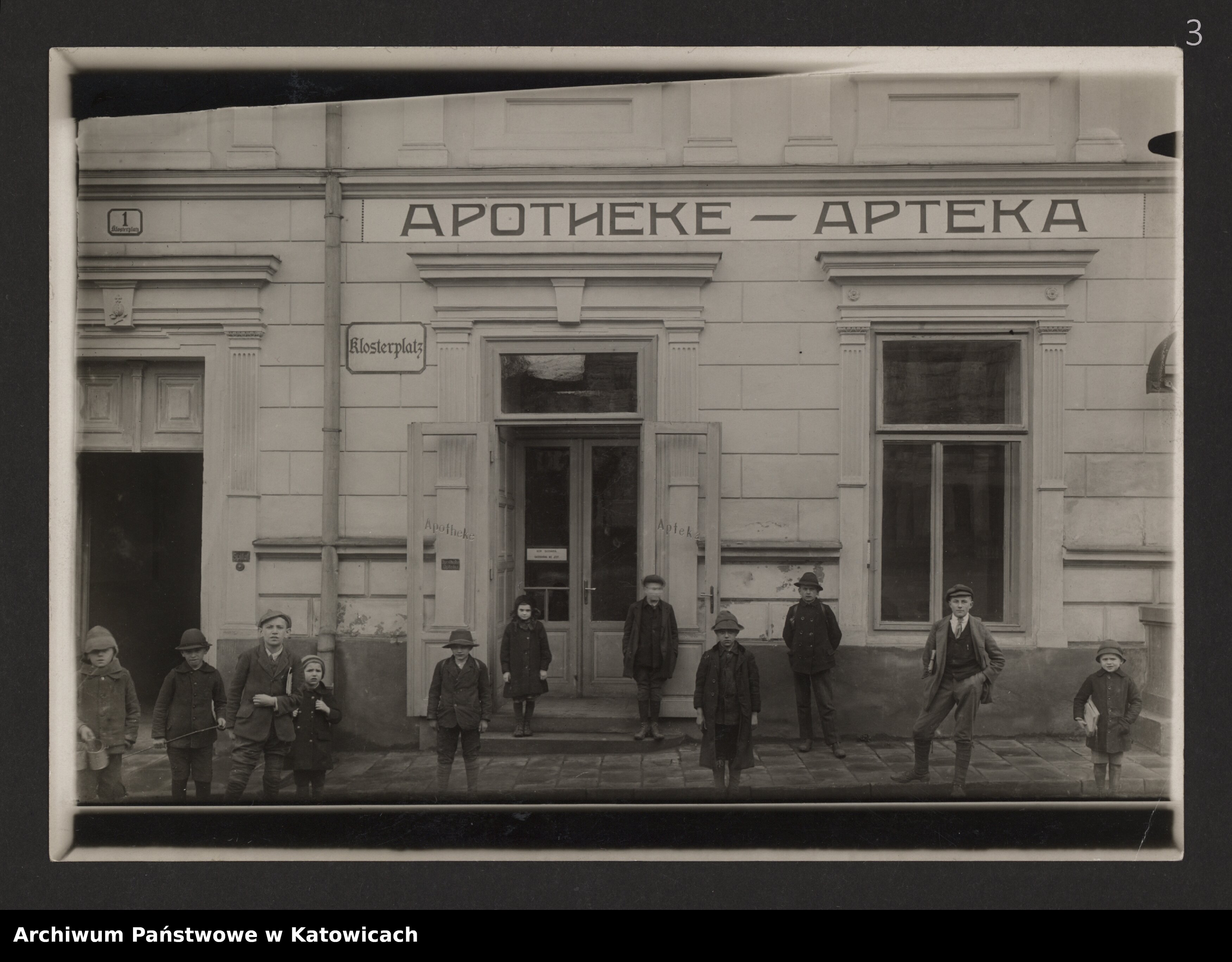 Zdjęcie ukazuje dzieci stojąe przed wejściem do Apteki w Cieszynie. Nad witryną widnieje dwujęzyczny napis 