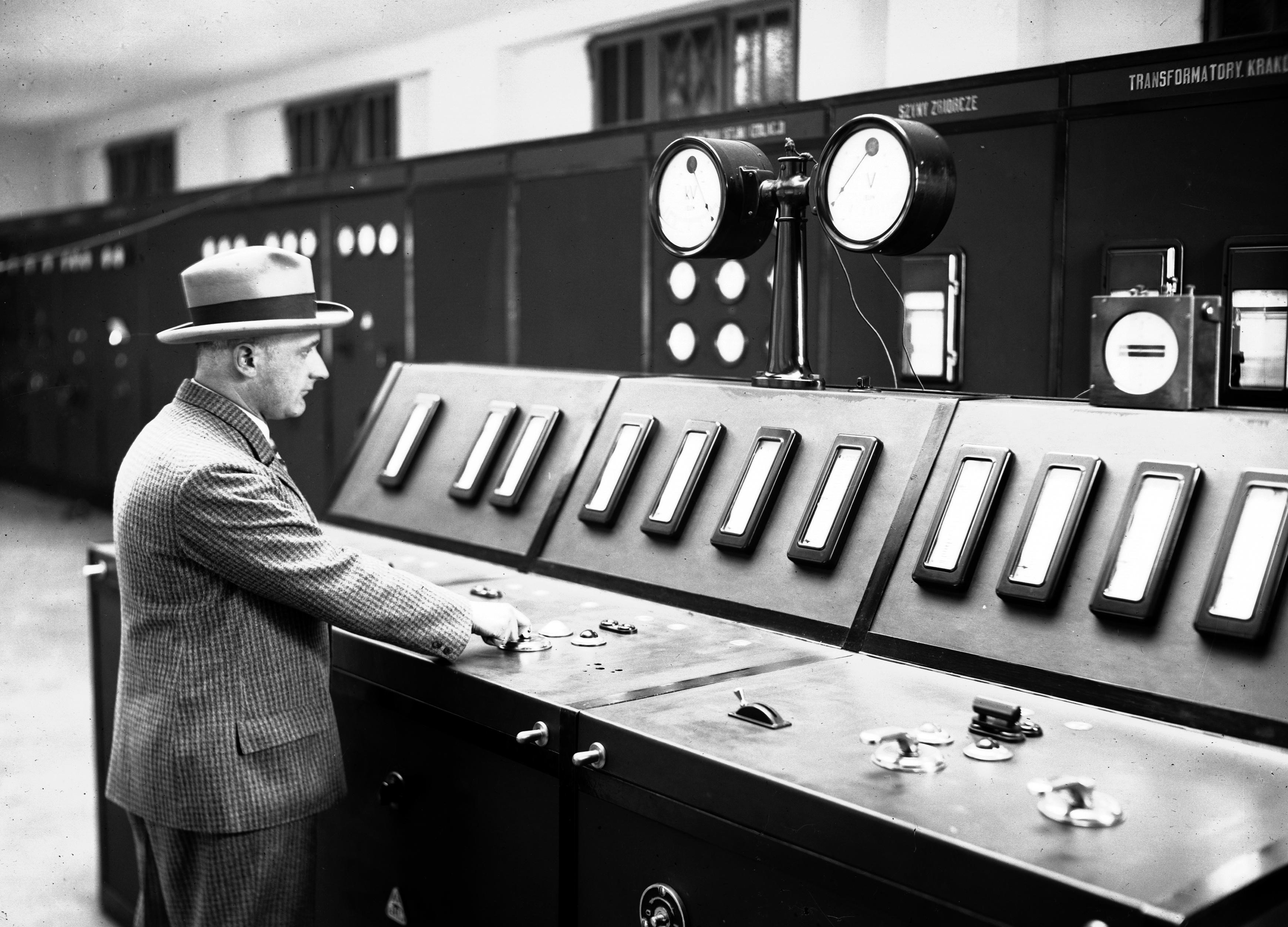 Czarno-biała fotografia przedstawia sterownię i rozdzielnię elektryczną elektrowni w Jaworznie przy pulpicie stoi inż. Stietz. 